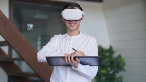 Smiling Asian Young Man in VR Headset Standing in Living Room Using Graphics Tablet alt