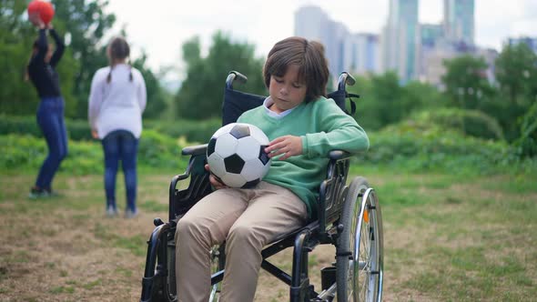 Portrait of Hopeless Caucasian Disabled Boy in Wheelchair Sitting with Ball Looking at Camera with alt