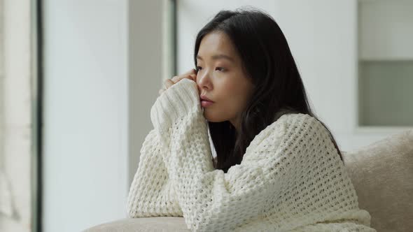 A Young Unhappy Asian Woman Sitting on the Couch with a Sad Face and Looking Out the Window alt