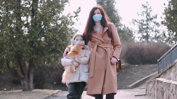 European Mother and Her Daughter in Masks Are in a Park with a Dog alt