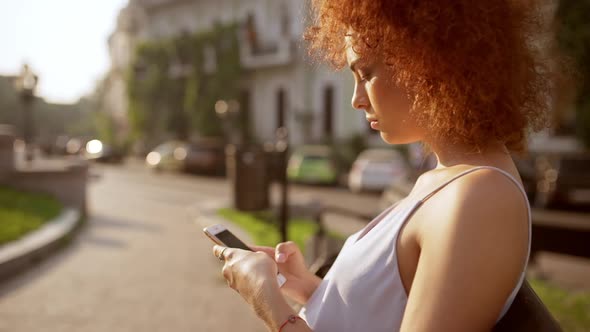 Attractive Foxy Girl Typing on Phone Sitting on Bench in City Park Slow Motion alt