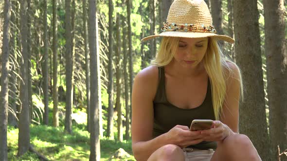 A Young Beautiful Woman Sits in a Forest on a Sunny Day and Works on a Smartphone alt