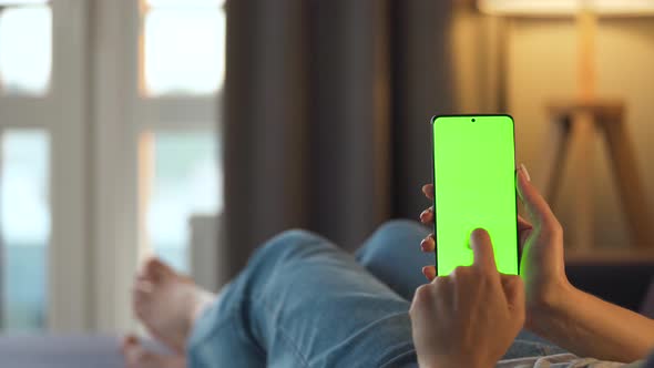 Woman at Home Lying on a Sofa and Using Smartphone with Green Mock-up Screen in Vertical Mode. Girl alt