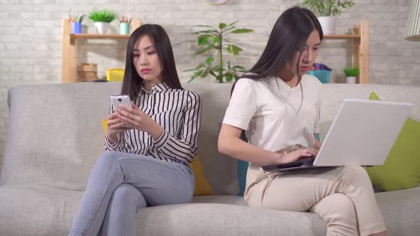 Two Young Asian Women Are Using Gadgets Sitting on the Sofa alt