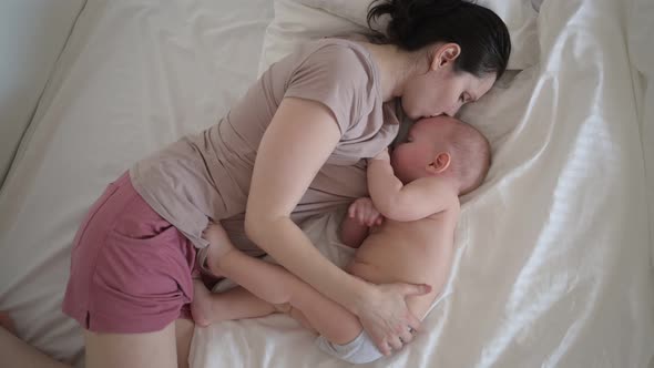 Young Mother Lying with Newborn Cute Infant Naked Baby Boy on Bed Holding Him on Arms Hugging and