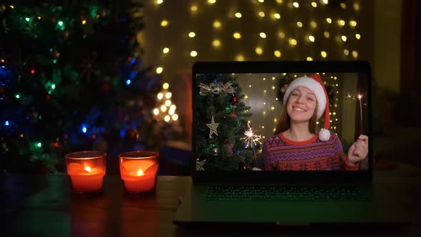 Happy Woman with Sparklers and Santa Hat Congratulates Relatives on Christmas Via Laptop Online