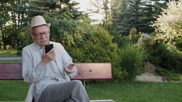 Senior Man Chatting on Web Camera Smiling and Gesturing When Resting in Park alt