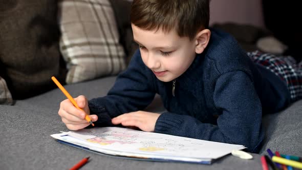 Little boy draws in coloring with colored pencils on sofa at home.