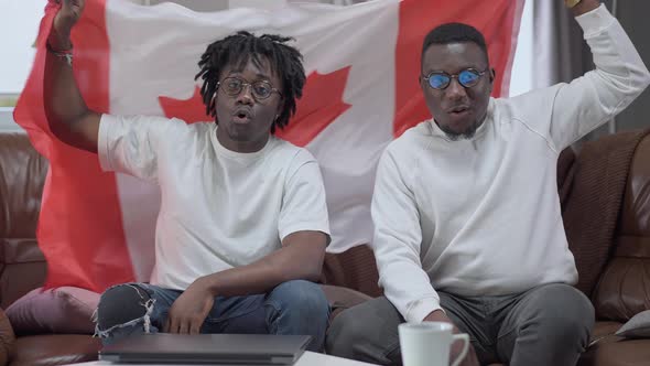 Portrait of Cheerful African American Men Shaking Canadian Flag Sitting Indoors on Couch Looking at alt