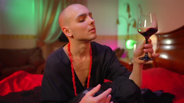 Confident Male Queer Shaking Red Wine in Glass Looking at Camera Sitting in Bedroom with Red Light alt