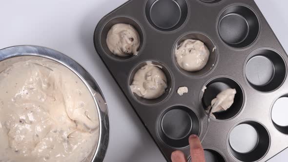 Batter being carefully scooped into a muffin pain. Top down view of stainless bowl and pan.