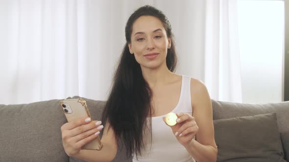 A Young Girl Holds a Bitcoin and a Phone in Her Hands While Sitting on the Couch at Home alt