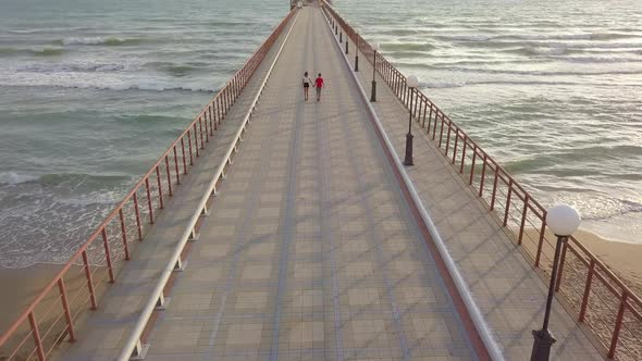 Aerial view of teen couple boy and girl walk by hand on pier in sea at sunset. Summer vacation, love