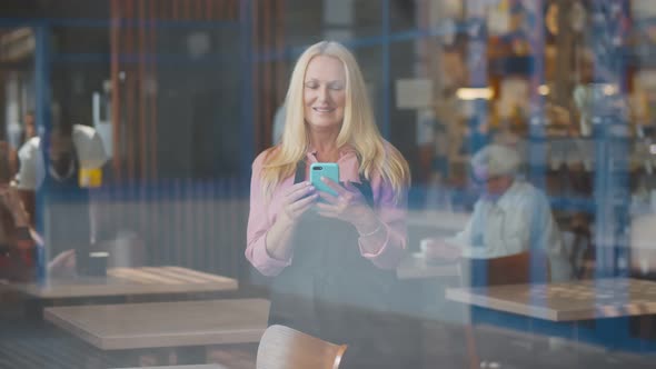 View Through Window of Smiling Aged Waitress in Apron Using Smartphone Standing in Cafe alt