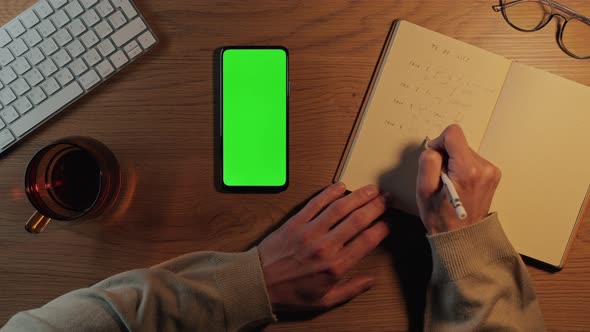 Man Scrolling on Green Screen Mobile and Writing in Planner alt