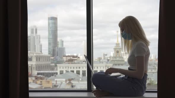 Freelancer Woman in Medical Mask Works at Home Using Laptop While Sitting on the Windowsill of a