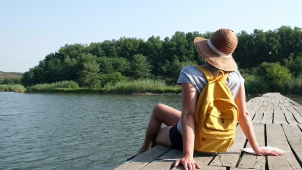 Traveler Hiker with Backpack Sitting on Wooden Bridge Across River in the Forest Resting alt