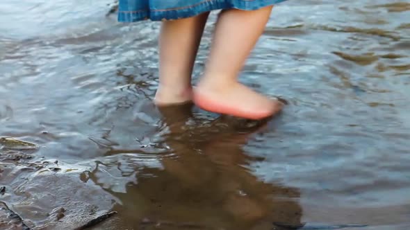 Legs of a Little Girl in a Denim Dress Walking on the Sand of a Riverbank in the Water in Summer