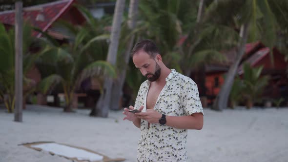 Traveler Man Using the Phone on the Beach Next to a Row of Beachfront Villas in the Evening alt