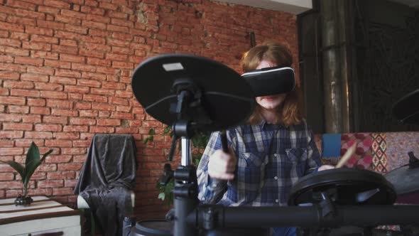 Young Redhead Man Musician Playing Electric Drums in Virtual Reality Glasses at Home Slow Motion alt