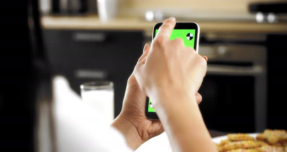 Woman Holds a Smartphone with a Green Screen in Her Hands Sitting in the Kitchen