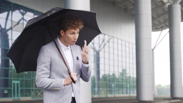 Serious Looking Businessman in Suit Standing with Umbrella Under Rain and Waiting for His Driver