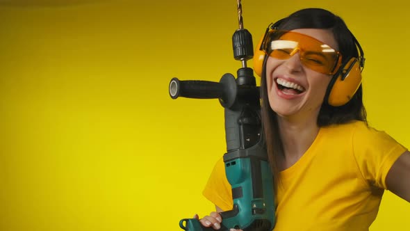 A Cheerful Woman in Protective Headphones is Holding a Drill in Her Hand alt