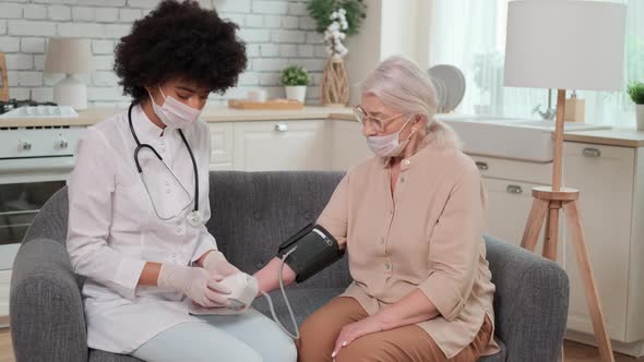 Afro American Woman Doctor in Mask Checks Blood Pressure Senier Woman at Home Sitting on Couch alt