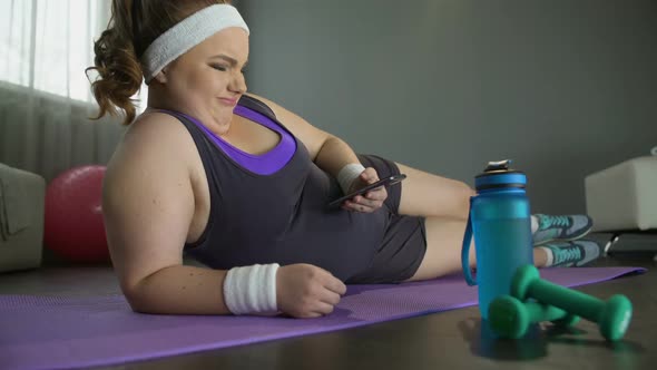 Fat Lazy Woman Lying on Floor With Smartphone in Hands Instead of Training alt