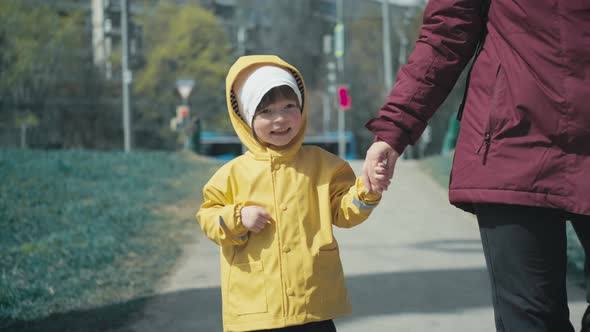Child in Yellow Raincoat with Hood Walks Through City and Holds Hand of His Mom