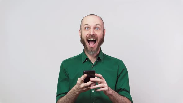 Happy bearded man looking surprised using mobile phone over white background.