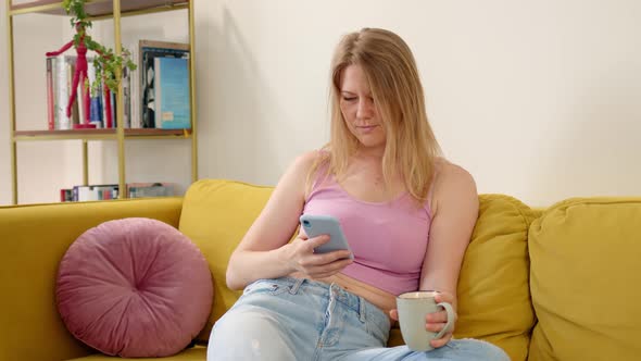 Nice Woman in Pink Tank Top and Blue Jeans Sits on Yellow Sofa and Drinks Hot Tea or Coffee From alt