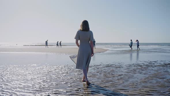 Woman Walks on Beach with Watery Pools Leading to the Ocean on a Windy Sunny Day alt
