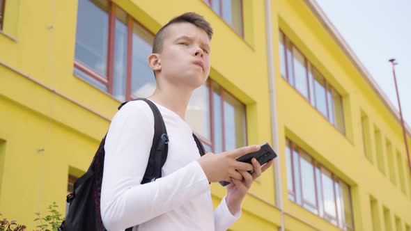 A Caucasian Teenage Boy Works on a Smartphone  View From Below  a School in the Background