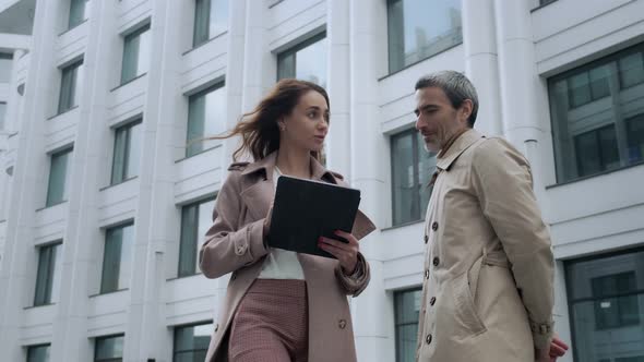 A beautiful girl with a tablet approaches the head of a man standing near office buildings alt
