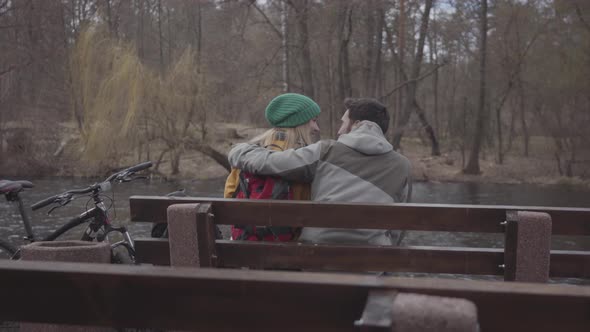 Back View Young Couple of Tourists with Backpacks Sitting on the Bench in the Spring Park alt