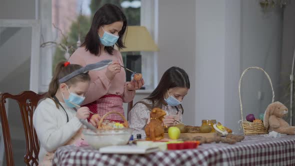 Focused Caucasian Woman and Girls in Coronavirus Face Masks Coloring Eggs on Easter Indoors alt