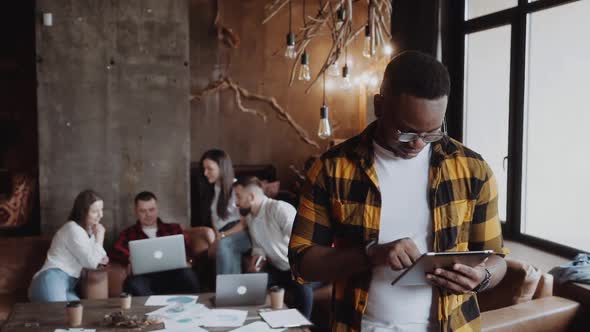 Afro American Man in Casual Uses a Tablet in a Modern Loft Office Colleagues at the Background alt