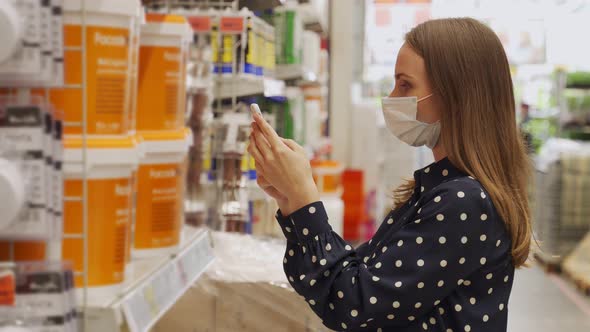Woman Wearing a Protective Mask Picks Up Paint at a Hardware Store and Uses a Smartphone alt