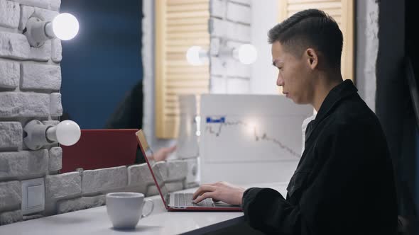 Focused Asian Man Typing on Laptop Keyboard Analyzing Graph in Home Office at Night alt