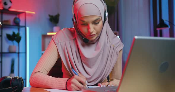 Arabic Woman in Hijab Wears Wireless Earphones Listening Online Presentation on Computer at Home alt