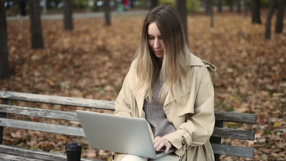 Young Beautiful Woman Works at a Laptop Sitting on a Park Bench with Coffee alt