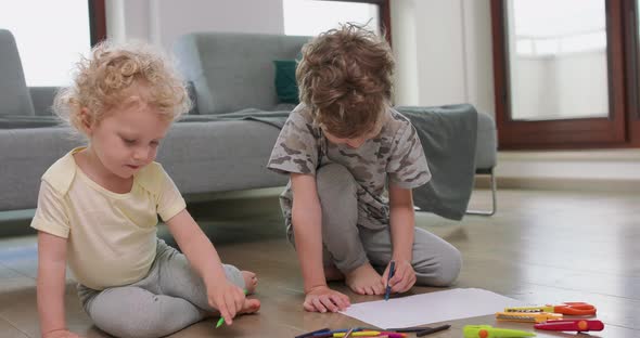 A Little Boy and a Little Girl are Drawing with Pencils on on the White Paper on the Floor Looking alt
