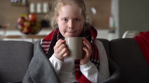 Portrait of a Sick Child in a Scarf and Plaid with a Cup of Hot Tea on the Sofa in the Apartment