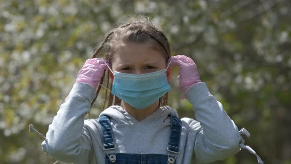 A Little Girl in Rubber Gloves Puts on a Medical Mask Against the Background of a Flowering Tree