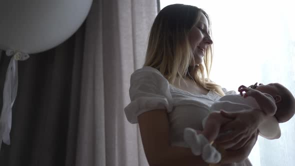 Portrait of Loving Caucasian Woman with Newborn Baby in Hands Standing at Window Indoors Admiring alt