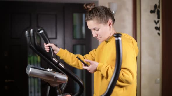 girl in a yellow sweatshirt idly trains on an ellipsoid and looks at the phone alt