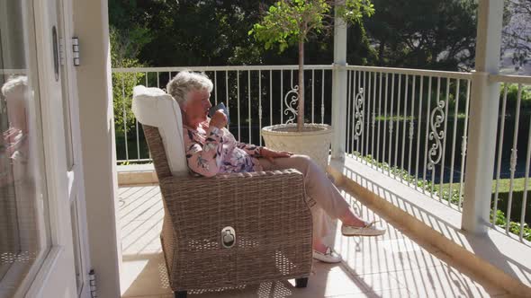 Caucasian senior woman drinking coffee while sitting on a chair in the balcony at home alt