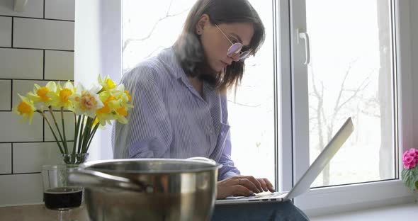 Young brown haired 20s woman cooking dinner or lunch and sitting near window with laptop.  alt