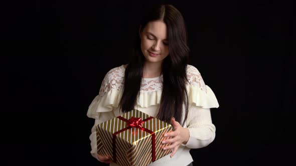 Young Woman with a Gift Box on Black Background. Gift Box with White Ribbon for Happy New Year alt